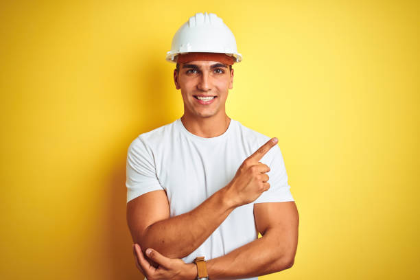 Young handsome man wearing construction helmet over yellow isolated background with a big smile on face, pointing with hand and finger to the side looking at the camera. Young handsome man wearing construction helmet over yellow isolated background with a big smile on face, pointing with hand and finger to the side looking at the camera. sign human hand pointing manual worker stock pictures, royalty-free photos & images