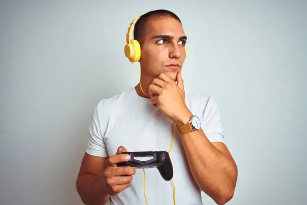 Young handsome man playing videogames using headphones over white isolated background serious face thinking about question, very confused idea