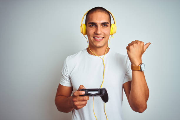istock Young handsome man playing videogames using headphones over white isolated background pointing and showing with thumb up to the side with happy face smiling 1187208359