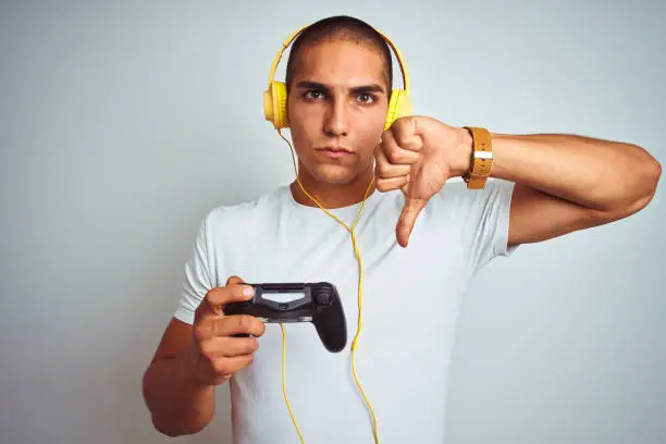 Young handsome man playing videogames using headphones over white isolated background with angry face, negative sign showing dislike with thumbs down, rejection concept