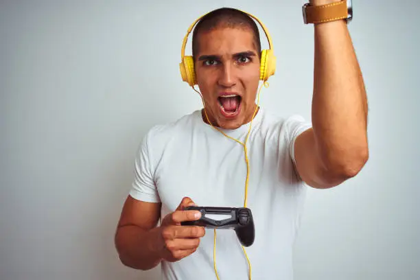 Young handsome man playing videogames using headphones over white isolated background annoyed and frustrated shouting with anger, crazy and yelling with raised hand, anger concept