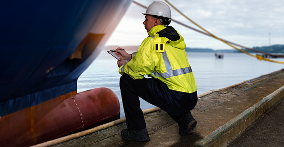 Ship supervisor engineer inspector stands at the dockside in a port. Wearing safety helmet and yellow vest. Cargo shipping industry. Protection and idemnity concept.