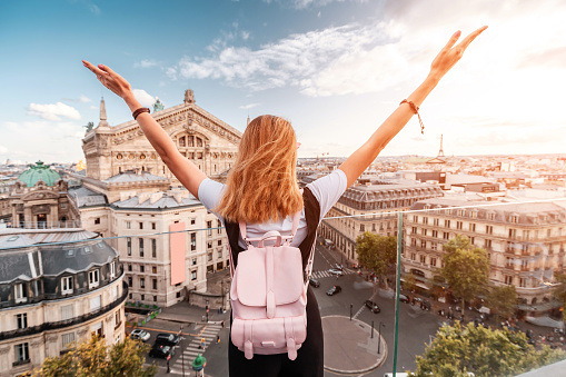 Happy asian woman with backpack standing on a roof top and enjoys great view over Parisian skyline at sunset. Travel and education in France