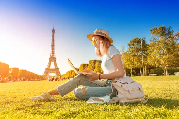 Photo of An Asian girl works on a laptop sitting on the grass on the Champ de Mars in Paris overlooking the Eiffel tower at sunset. Concept of remote work and freelancing