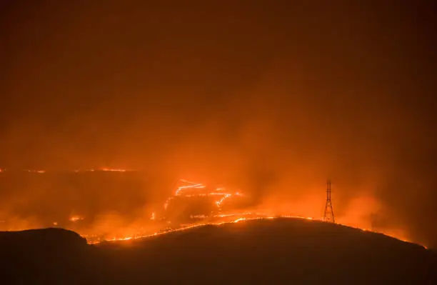 Photo of A massive wildfire burns its way in to Grand Coulee Washington at night. The fire and the city glow in the smoke night air. Silhouettes of the power line towers that connect the Grand Coulee Dam can be clearly seen.