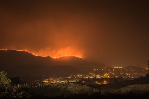 A massive wildfire burns its way in to Grand Coulee Washington at night. The fire and the city glow in the smoke night air. Silhouettes of the power line towers that connect the Grand Coulee Dam can be clearly seen. The Grass Valley Wildland fire was driven by strong wilds leading to an intense and swiftly moving fire that burned more than 75,000 acres. Firefighters worked into the night holding the wind driven wild fire from reaching any buildings in the town. un enorme incendio forestal se llega a grand coulee washington por la noche. el fuego y la ciudad brillan en el aire de la noche de humo. las siluetas de las torres de la línea eléctrica que conectan la presa grand coulee se pueden ver claramente.