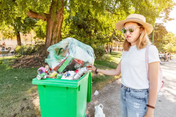The annoyed girl throws the garbage into the already filled garbage container. The concept of environmental pollution and inaction of the authorities