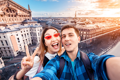 Loving couple Asian girl and European guy hug and take a selfie on the observation deck overlooking the Parisian landscape. Honeymoon in France