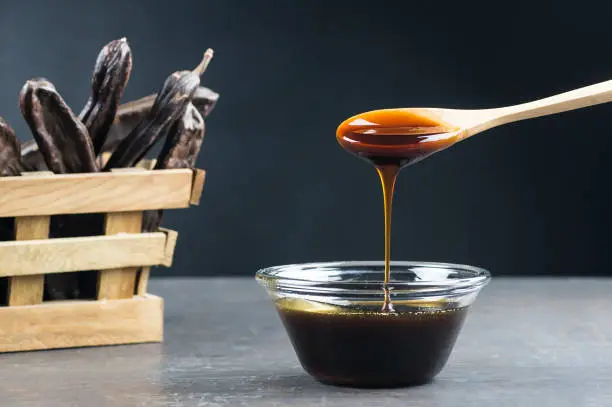 Photo of carob molasses in glass bowl and in wooden spoon and carob pods on rustic background, locust bean healthy food, Ceratonia siliqua ( harnup )