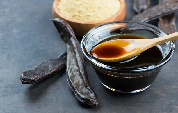 Photo of carob molasses in glass bowl and in wooden spoon and carob pods on rustic background, locust bean healthy food, Ceratonia siliqua ( harnup )