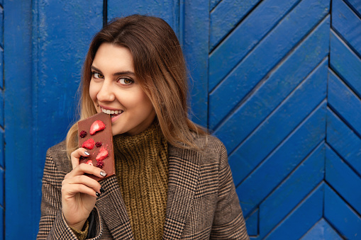 Winter. Woman with pleasure eating handmade chocolate with strawberries on blue wall background. Concept of sweet life