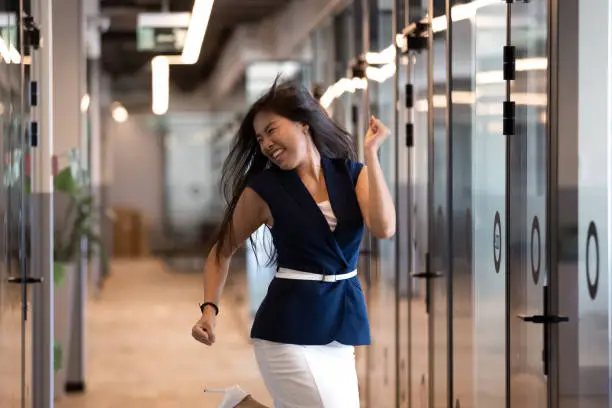 Photo of Excited Asian businesswoman celebrating success, dancing in office hallway