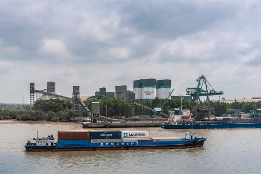Long Tau River, Vietnam - March 12, 2019: Nhon Trach area. Gemadept container river boat sails past INSEE Cement station with silos, conveyor belt, and its quay under dark cloudscape. Green belt and brown water.