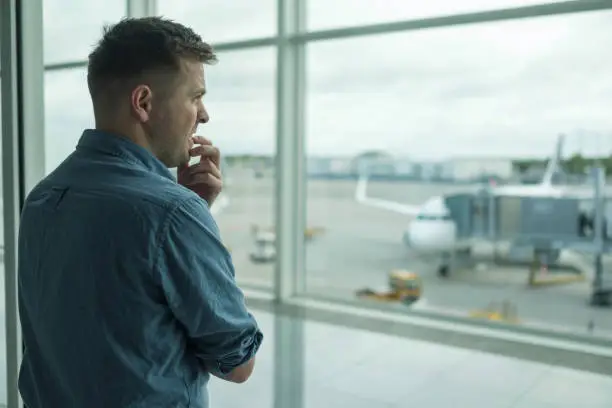 Photo of Caucasian young man is afraid to flight standing in terminal near window