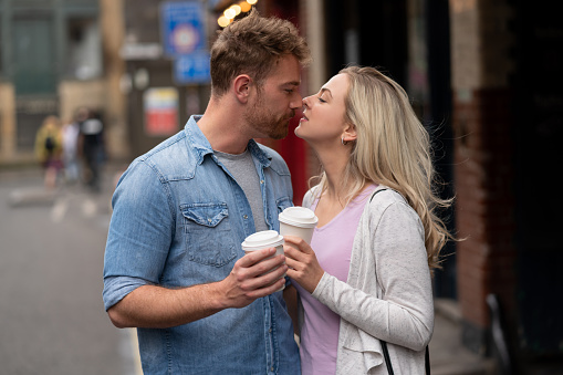 Portrait of a happy couple kissing goodbye on the street while drinking a cup of coffee