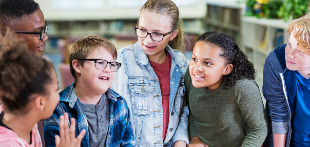 A multi-ethnic group of six elementary school students standing together in the library. They are 10 and 11 years old. The boy in the plaid shirt, wearing eyeglasses, has down syndrome.