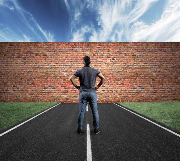 Young man, his back to the camera while stands in the middle of a road that runs directly into a large brick wall stock photo