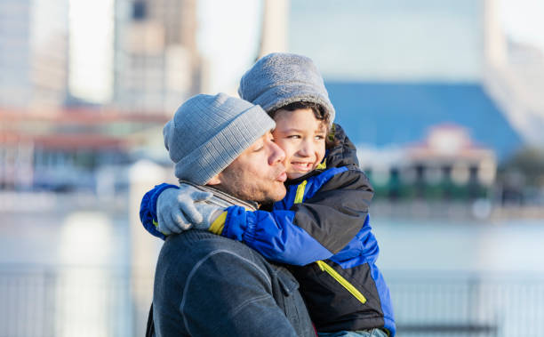A 6 year old Hispanic boy with his father on a city waterfront on a bright, sunny autumn or winter day. They are wearing warm clothing - jackets, knit hats, scarves and gloves. Dad is carrying his son in his arms, talking to him as they watch something.