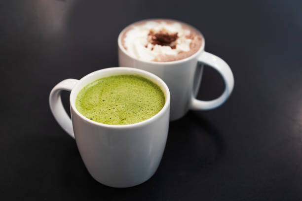 White mug of matcha green tea next to hot chocolate with selective focus - Freshly brewed healthy Japanese latte drink full of antioxidents and health benefits - cafe, winter and coffee shop concept