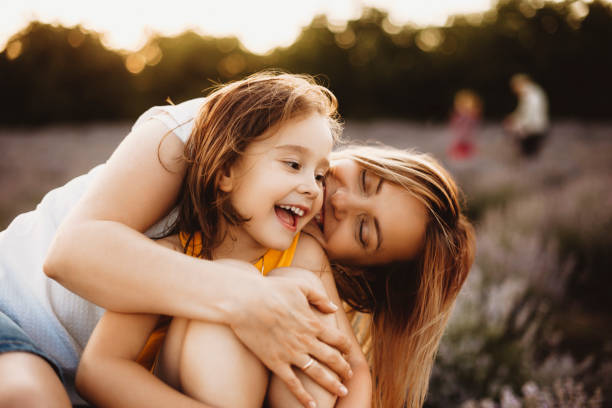 夕焼けに対する花のフィールドで屋外で彼女の若い母親に抱きしめながら笑う美しい女の子。 - lavender coloured ストックフォトと画像