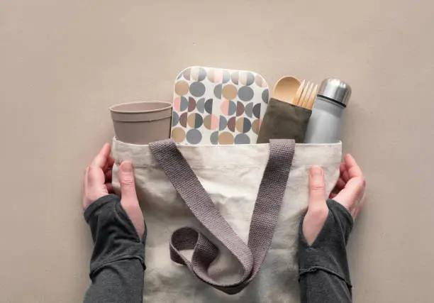 Creative top view, zero waste packed lunch kit in canvas bag. Hands holding bag with takeaway lunch box, bundle with bamboo cutlery, reusable box and coffee-to-go cup. Flat lay on brown craft paper.