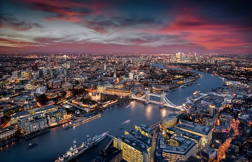 Aerial view of illuminated London, UK, during evening time featuring the Tower Bridge, Thames river and the modern skyscrapers of Canary Wharf