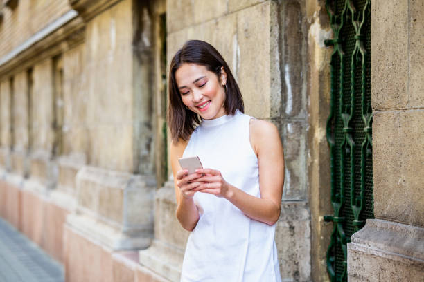 istock Beautiful asian woman using an application to send an sms message in her smartphone device. Happy young Asian woman with smartphone standing in the street 1187308831