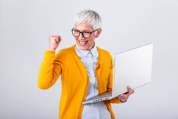 Portrait of a cheerful mature woman with a laptop computer and celebrating success isolated over gray background. Senior lady watching celebrating online bid bet win or great result victory concept Portrait of a cheerful mature woman with a laptop computer and celebrating success isolated over gray background. Senior lady watching celebrating online bid bet win or great result victory concept ecstasy stock pictures, royalty-free photos & images