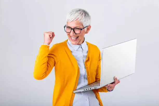 Photo of Portrait of a cheerful mature woman with a laptop computer and celebrating success isolated over gray background. Senior lady watching celebrating online bid bet win or great result victory concept