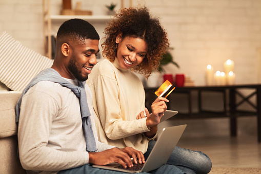 Online shopping. Happy african-american couple looking at laptop and buying with credit card at home, free space