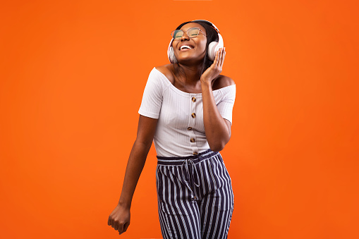 Music App. Cheerful Black Girl In Headphones Listening To Favourite Song And Dancing Over Orange Studio Background.