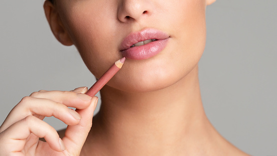 Beauty concept. Woman drawing lips with nude pink lipliner over grey background, crop