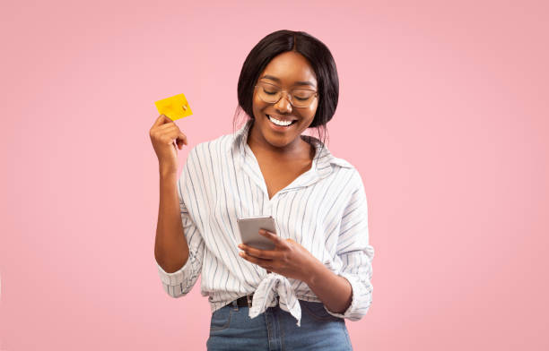 Afro Girl Using Cellphone And Credit Card Standing, Studio Shot Mobile Banking. Afro Girl Using Cellphone And Credit Card Standing Over Pink Background. Studio Shot credit card women internet currency stock pictures, royalty-free photos & images