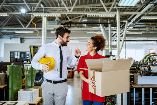 istock Friendly caucasian bearded director holding helmet under armpit walking with his employee and talking. Employee holding box and looking at him. Printing shop interior. 1187322490