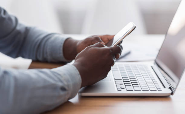 Unrecognizable afro businessman messaging on smartphone and working with laptop