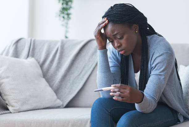 Depressed black girl holding pregnancy test, upset with positive result Unintended pregnancy. Depressed african american woman upset with positive test results, sitting on couch at home with sad face expression unwanted pregnancy stock pictures, royalty-free photos & images