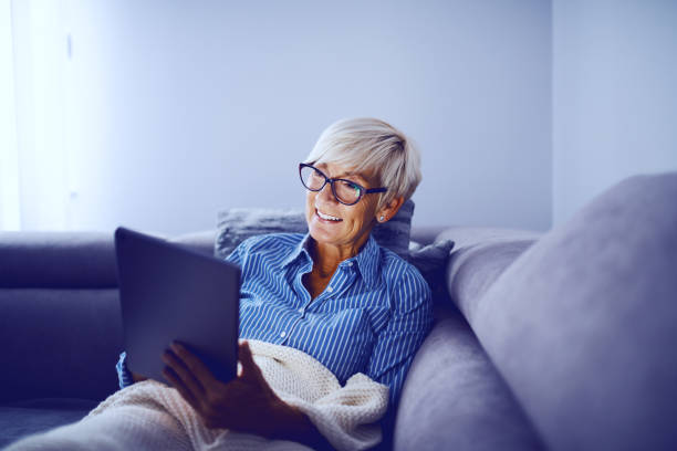 attraente donna anziana bionda sorridente caucasica seduta sul divano in salotto, usando il tablet e godendosi domenica pomeriggio. - ipad senior adult facebook sofa foto e immagini stock