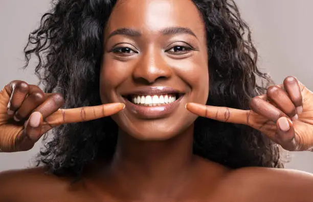 Teeth alignment and whitening concept. Happy african american girl pointing at her wide smile with two forefingers over grey background, closeup