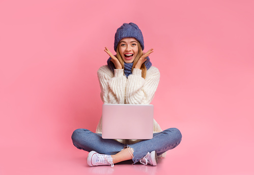 Big sales online. Beautiful young winter woman in knitted hat with laptop on her lap expressing happiness over pink background, sitting on floor