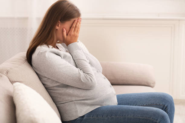 donna incinta che piange coprendo il viso con le mani sedute a casa - women sadness depression fear foto e immagini stock