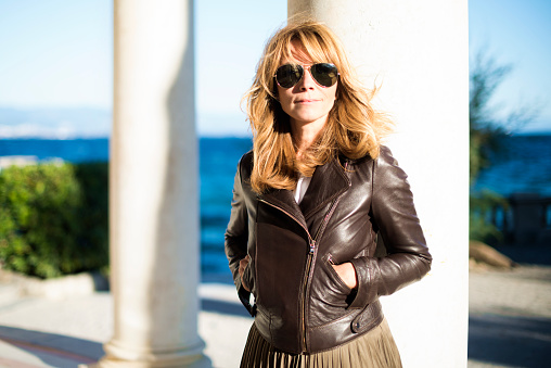 Close-up portrait shot of attractive mature woman wearing sunglasses and biker jacket while relaxing on terrace by the sea.