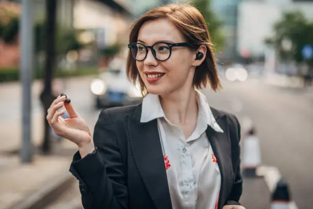 Businesswoman using wireless in ear headphones and mobile phone while walking on the street