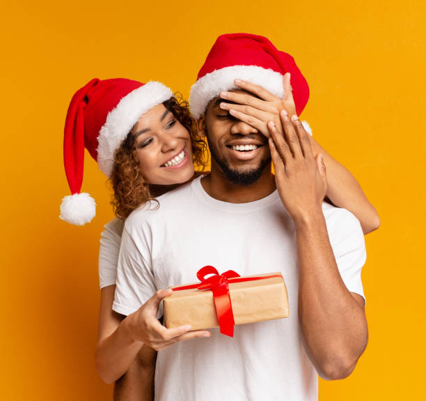 Black millennial couple with Christmas gift on orange background Loving african-american woman giving surprise gift to her boyfriend, close his eyes, orange background gift lounge stock pictures, royalty-free photos & images