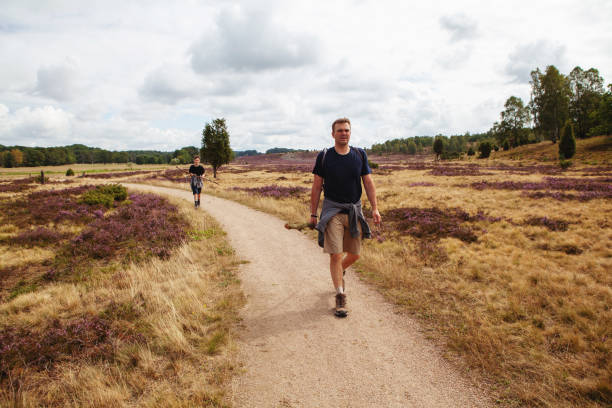 Father and his teenage son in shorts and t-shirts walking on the grassland in a nature reserve in Luneburger Heath among the heather plants on their hiking trip - being together, being active and healthy. Luneburg Heath, Germany - August 16, 2019: a man and his teenager boy have a stroll in the meadows of Lower Saxony in summer with their backpacks on. lüneburg heath stock pictures, royalty-free photos & images