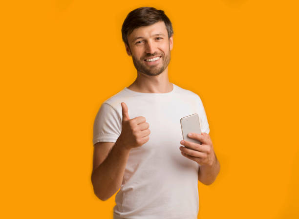 istock Guy Holding Phone Gesturing Thumbs-Up Standing On Yellow Background 1187344942
