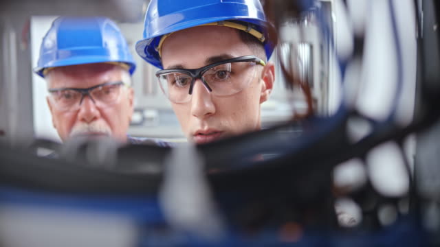 DS Face of a young male apprentice connecting wires in the electrical panel under supervision of a senior electrician