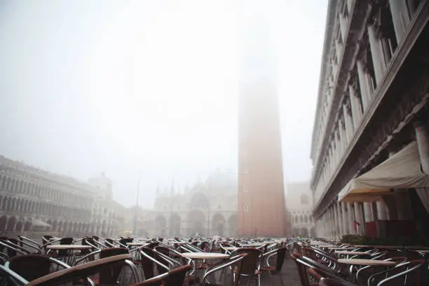 Venice. Italy - October 28, 2019: empty cafe tables and Piazza San Marco on a foggy morning just after the dawn with Campanile lost in mist and everything blurry and out of focus.