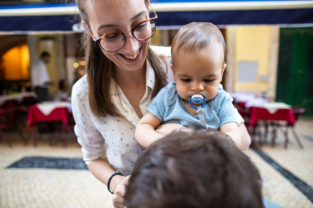 Young couple having fun with baby boy at sidewalk cafe, mother holding baby with pacifier in his mouth