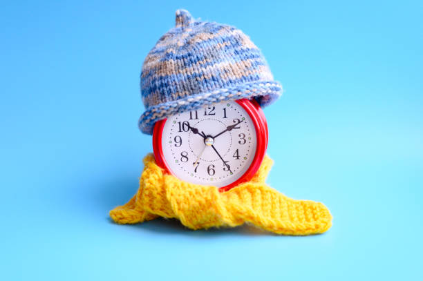 istock red round alarm clock in knitted wool blue hat and yellow scarf centered on a blue background. winter time concept. winter season. cozy and warm. analog time 10:10. space for text 1187365581
