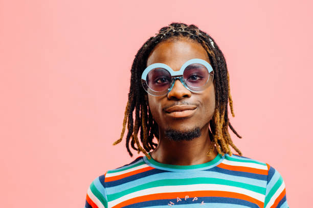 verticale d'un jeune homme de sourire dans la chemise rayée avec le signe ' heureux' et de grandes lunettes de soleil, restant devant un fond rose - men fashion model cool glasses photos et images de collection